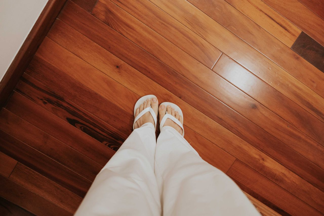 View of person wearing flip-flops and white pants standing on a wooden floor.